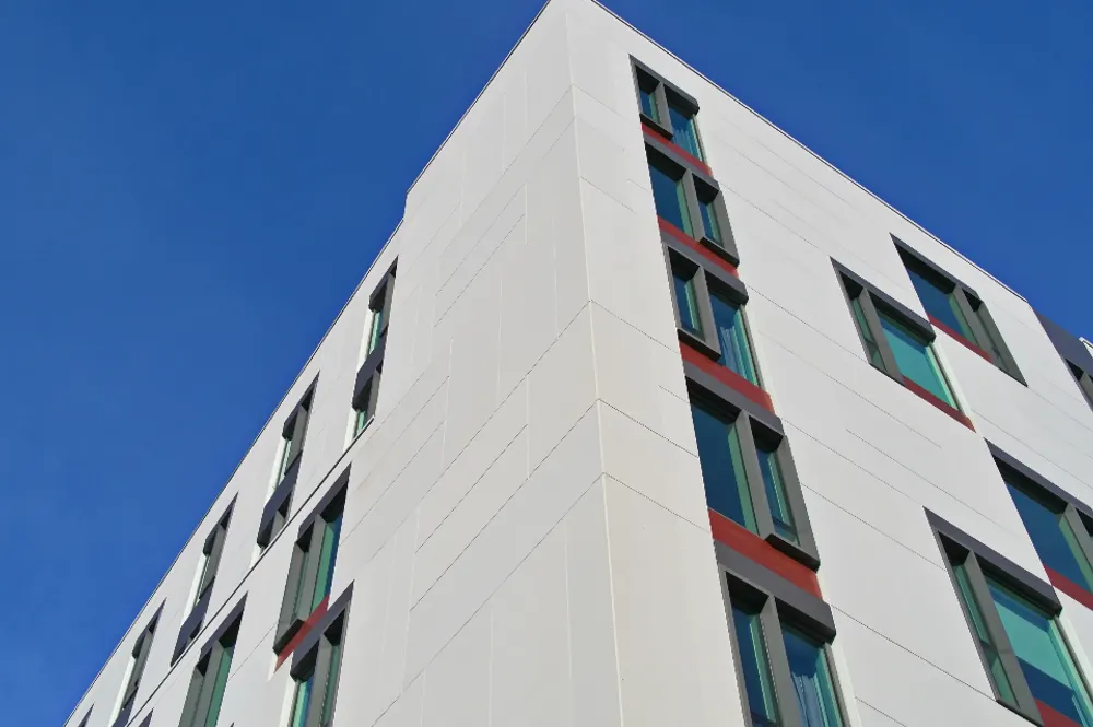 The corner of a white-clad hospital building with tall narrow windows, against a clear blue sky.
